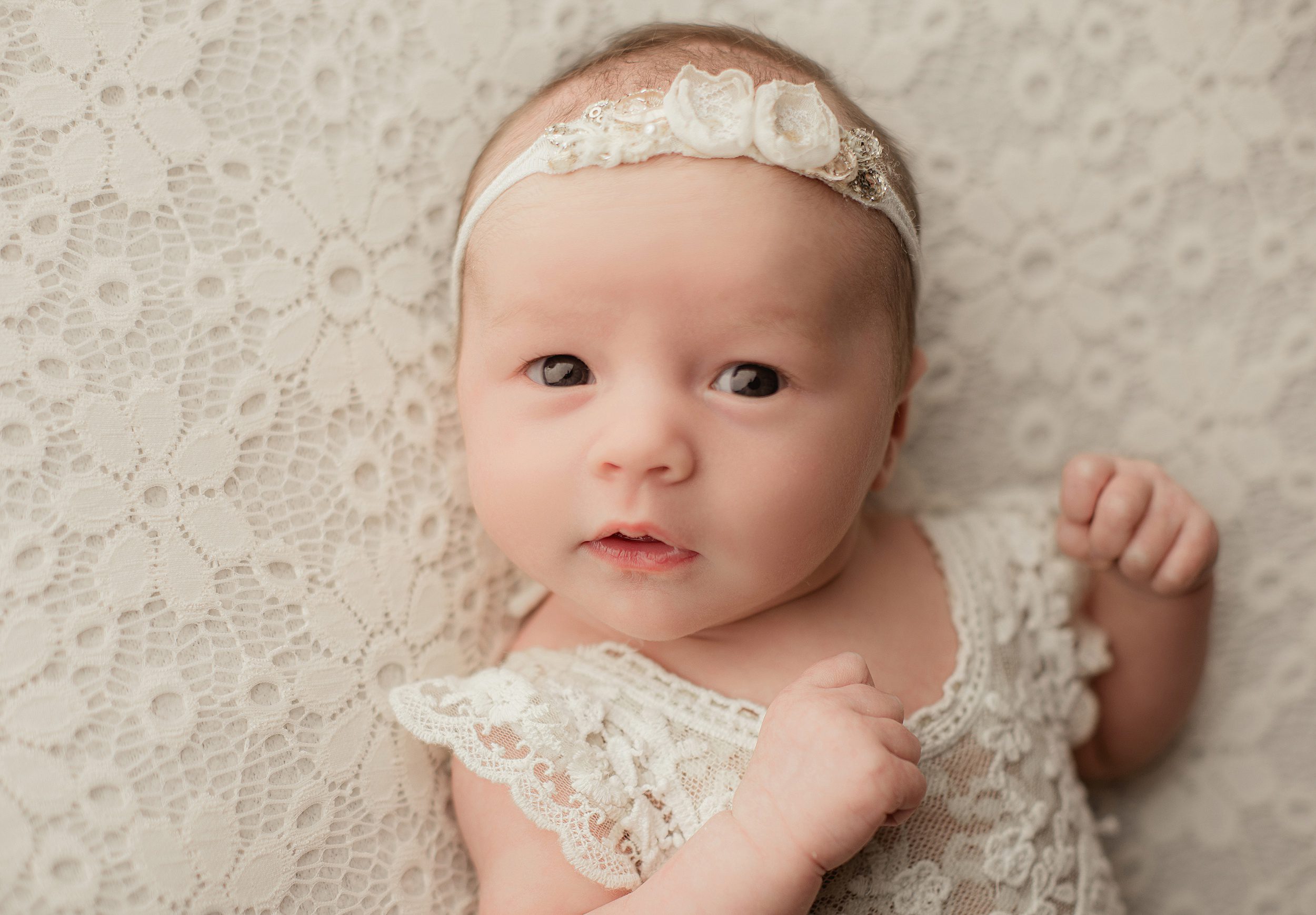 A newborn baby girl lays on her back in a lace dress and white floral headband