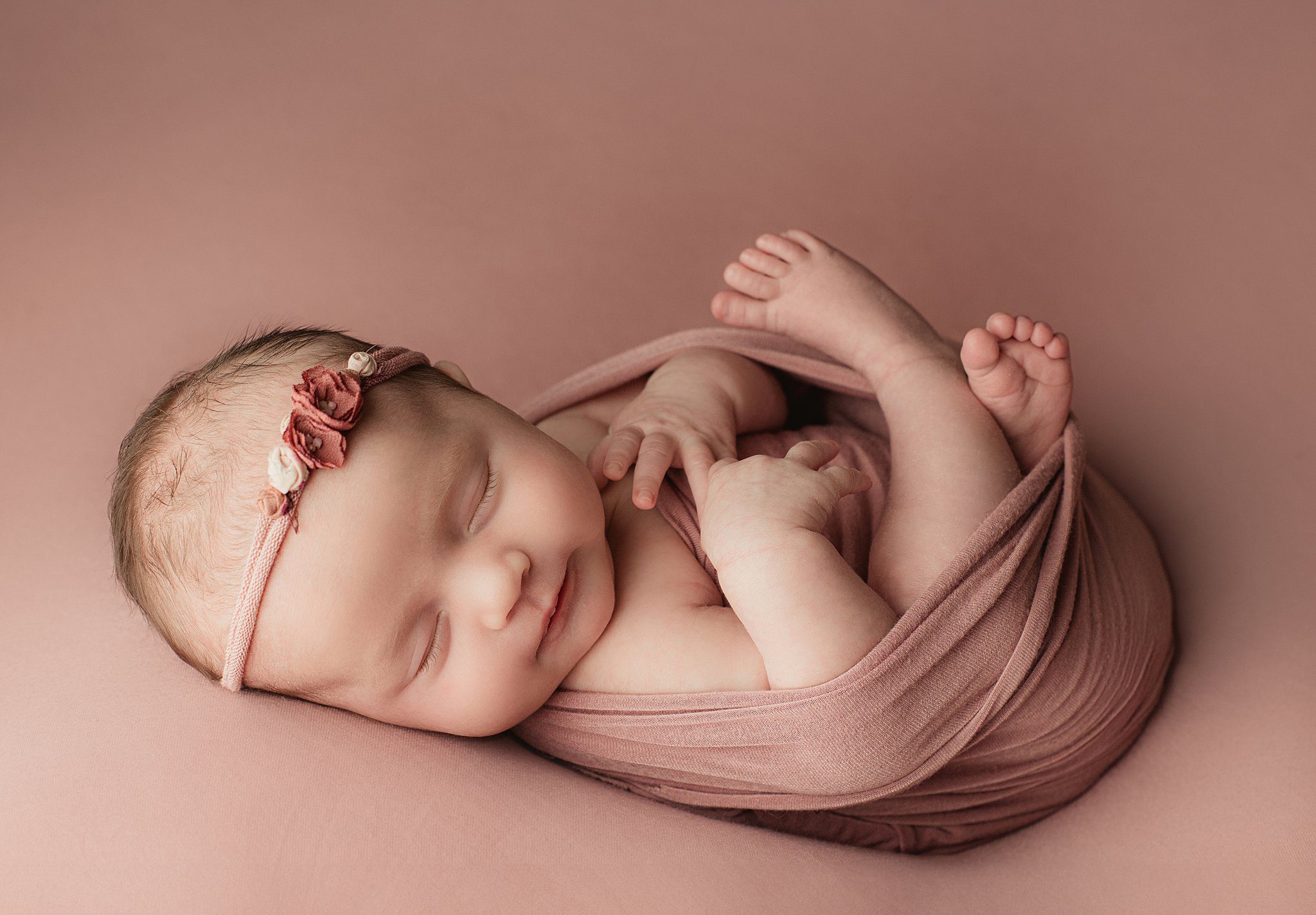 A newborn baby sleeps in a pink swaddle with legs and arms out