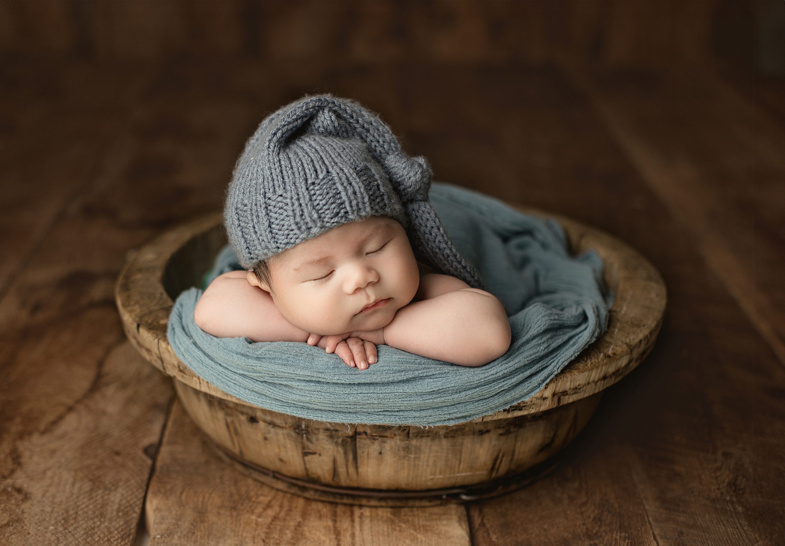 A newborn baby sleeps in a wooden bowl on a wood floor in a studio