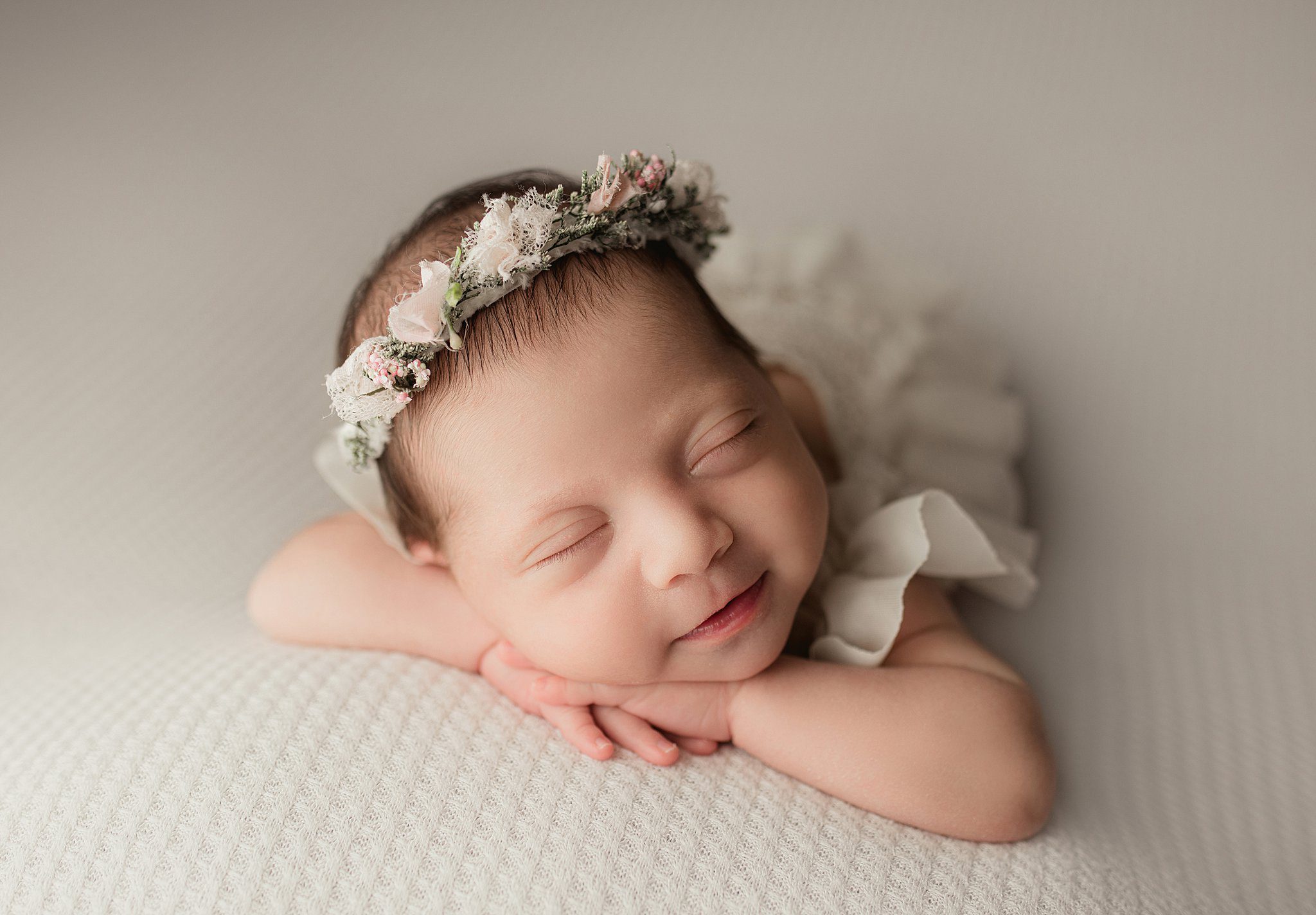 A newborn baby smiles in her sleep on a white bed in a white dress and headband
