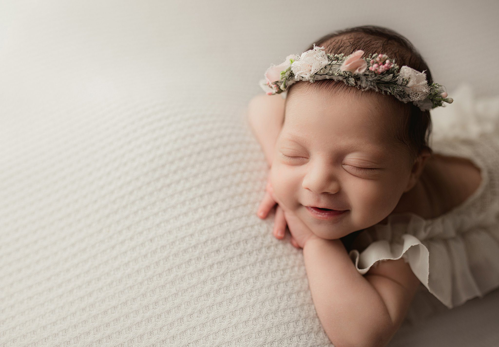 A newborn baby sleeps in a white dress and floral headband with a smile