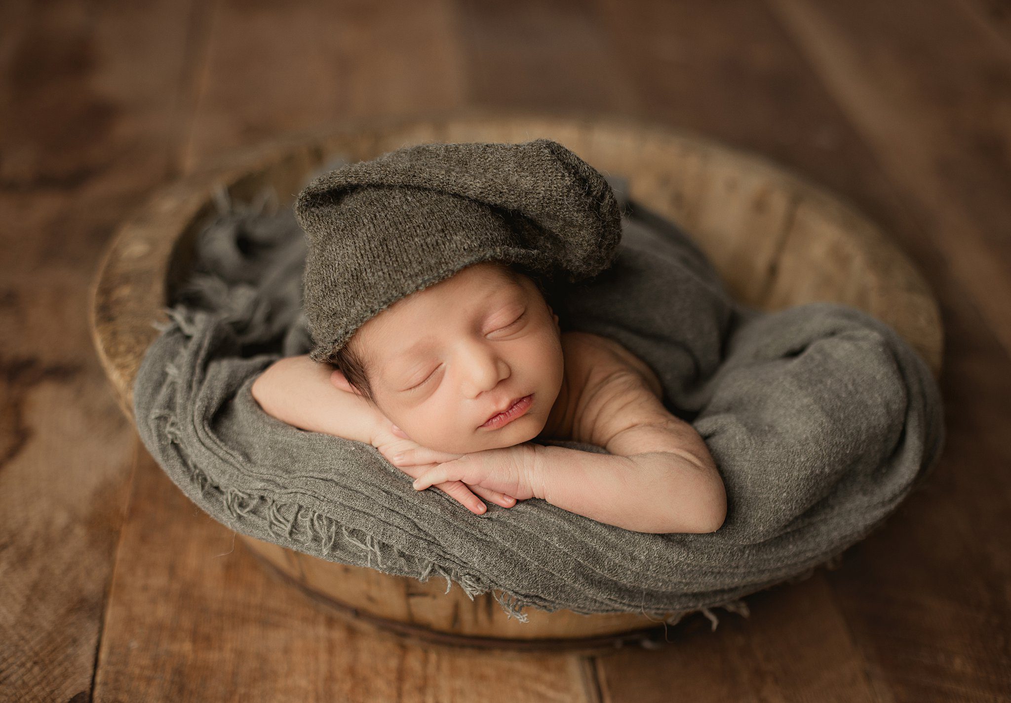 A newborn baby sleeps in a wooden bowl wearing a knit night cap