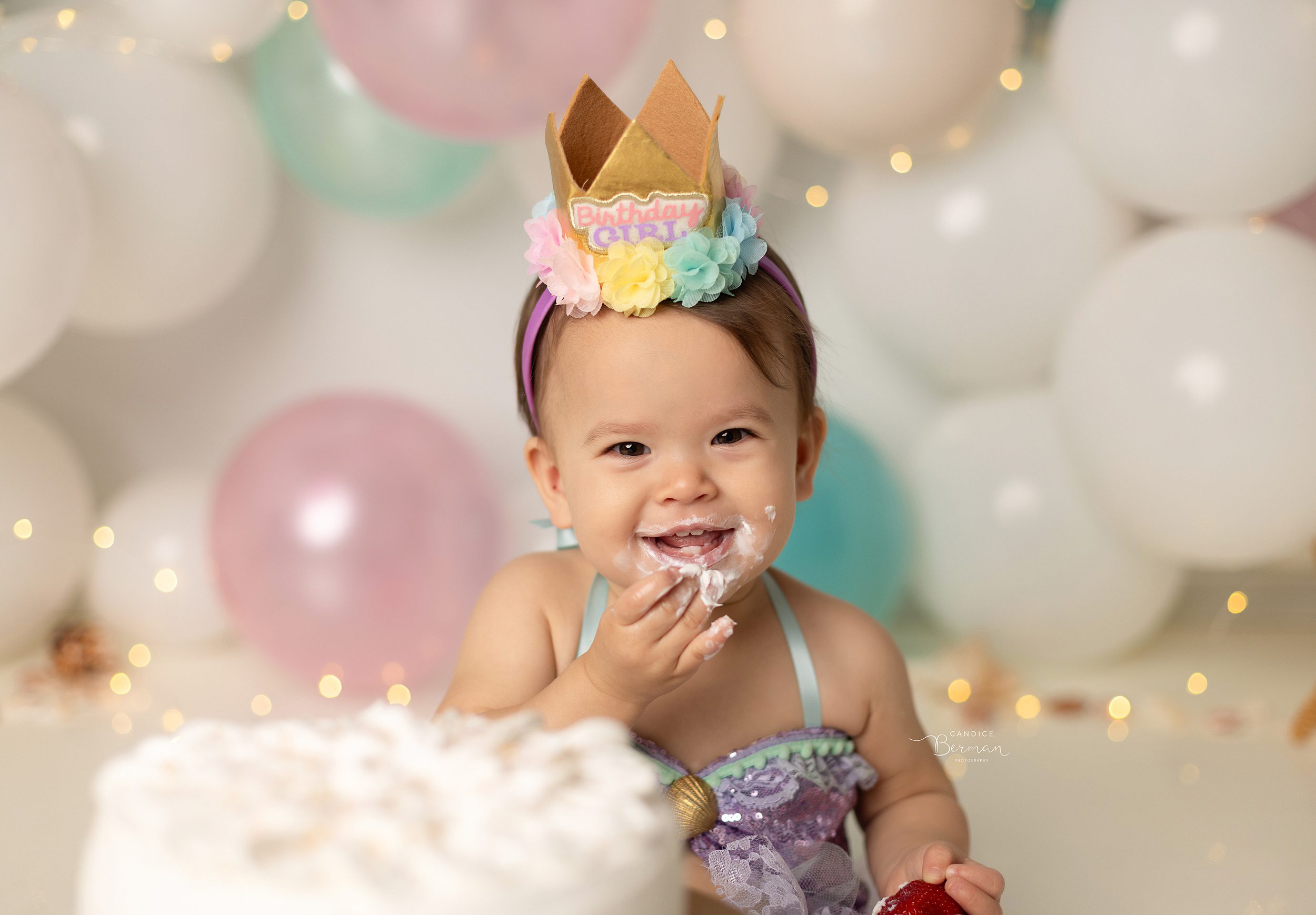 A toddler girl sits in a studio filled with balloons smashing a cake into her face before meeting orange county babysitters
