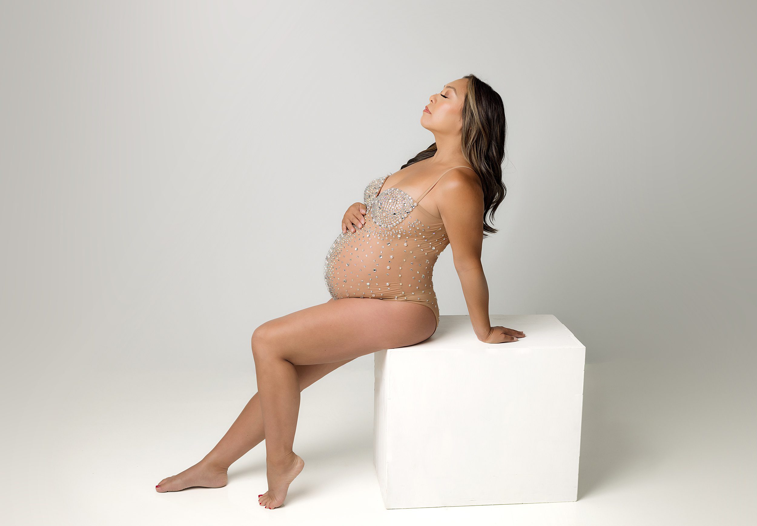 A mother to be sits on a white cube in a studio in a nude leotard after meeting with orange county midwives