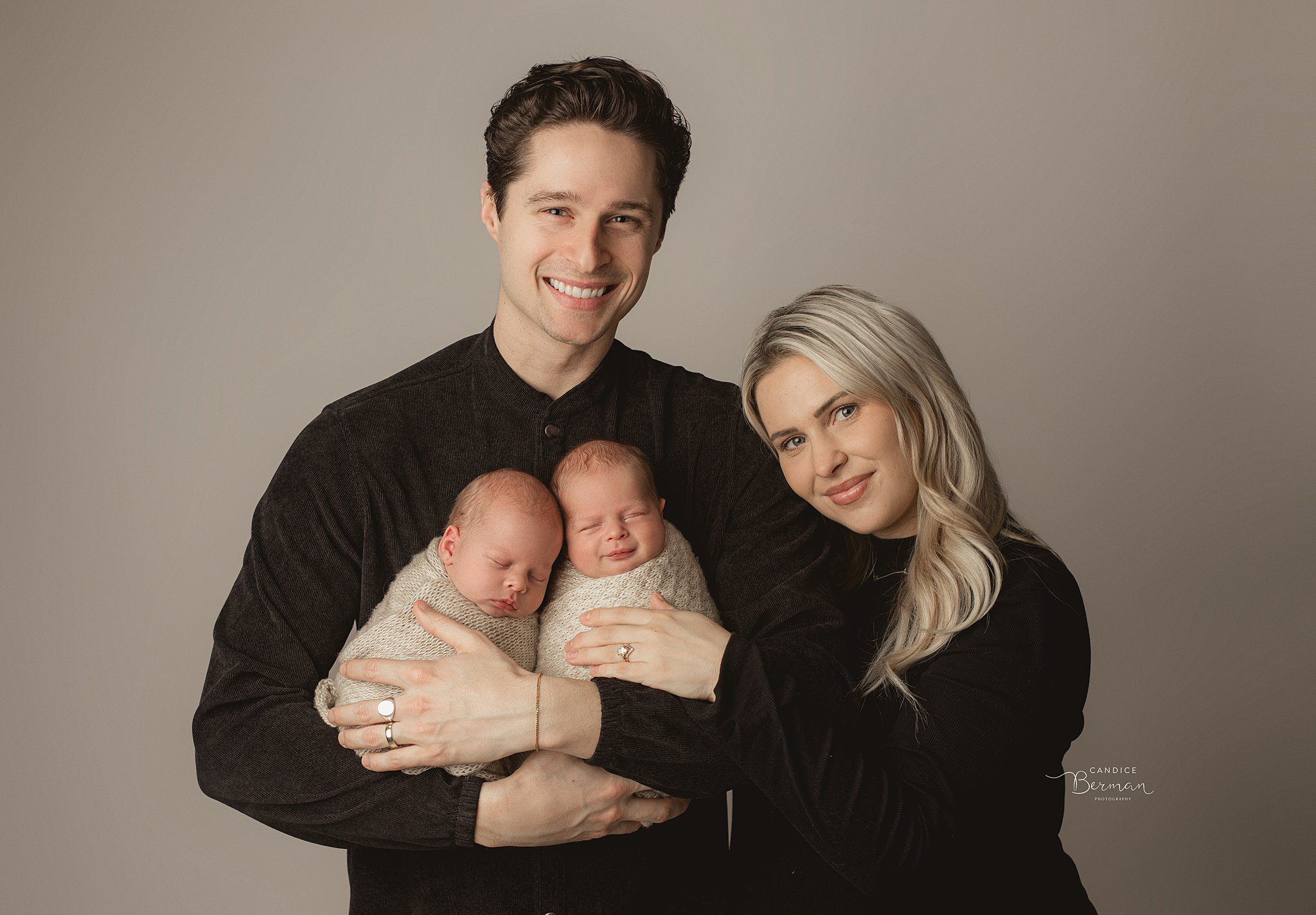 Happy new parents in black stand in a studio cradling their twin newborn babies as they sleep thanks to an orange county pediatric dentists