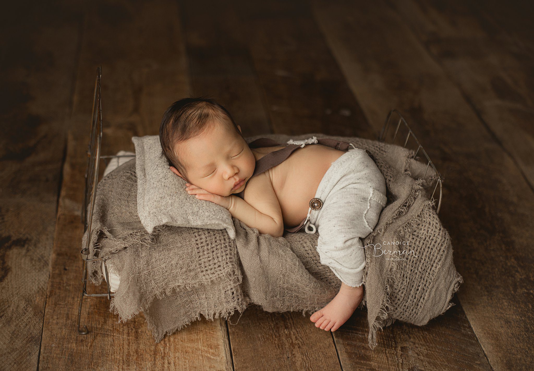 A newbown baby in grey pants and suspenders sleeps on a wire frame bed on hardwood floor in a studio