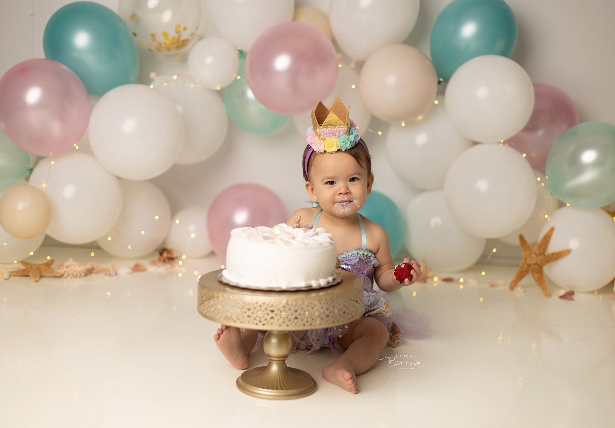 A toddler girl in a purple dress and birthday crown sits in a studio smashing a cake all over her face surrounded by balloons
